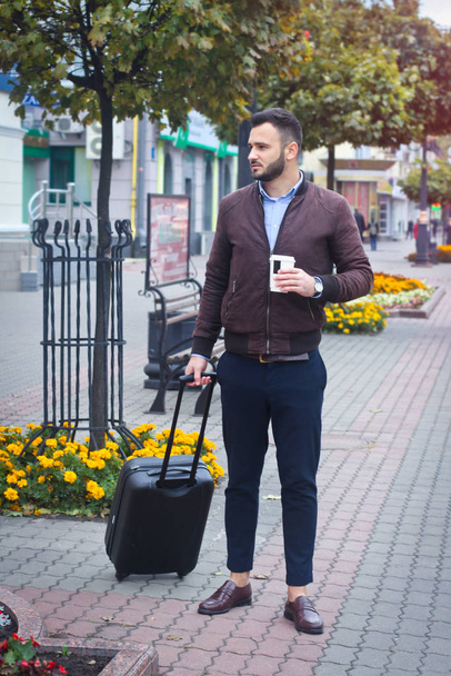 Handsome man with a suitcase for tourism. Exit the house for travel. Fashionably dressed guy and coffee. Stock Photo. - Zdjęcie, obraz