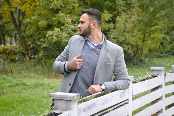 Πορτρέτο ενός σέξι άντρα ντυμένου με κοστούμι. Μοντέρνος τύπος στην φθινοπωρινή φύση κοντά σε ένα λευκό ξύλινο φράχτη. Ο επιχειρηματίας χαλαρώνει. Φωτογραφία Skok για σχεδιασμό - Φωτογραφία, εικόνα