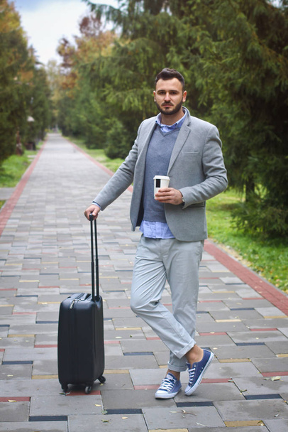 Fashionably dressed guy is traveling with luggage in the background of a road and trees. The man hurries on his flight and pours coffee, tea. Stock photo for design - Foto, Imagem