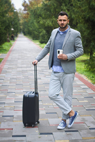 Fashionably dressed guy is traveling with luggage in the background of a road and trees. The man hurries on his flight and pours coffee, tea. Stock photo for design - Фото, изображение
