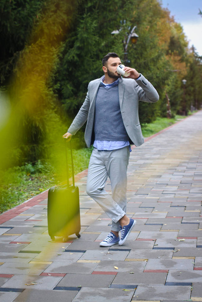 Modnie ubrany facet podróżuje z bagażem w tle drogi i drzew. Facet śpieszy się z lotem i nalewa kawę, herbatę. Zdjęcie w magazynie do projektu - Zdjęcie, obraz
