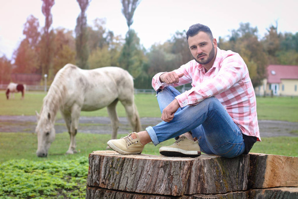 Young handsome guy Cowboy. farmer is sitting on a large stump on his ranch. Rural landscapes, countryside. Trees, field, farm. Stock photos for desig - Photo, image
