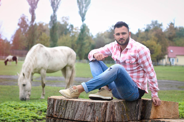 Young handsome guy Cowboy. farmer is sitting on a large stump on his ranch. Rural landscapes, countryside. Trees, field, farm. Stock photos for desig - Valokuva, kuva