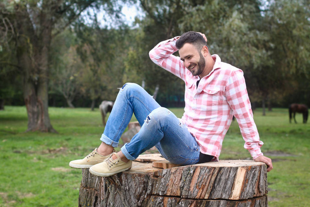 Young handsome guy Cowboy. farmer is sitting on a large stump on his ranch. Rural landscapes, countryside. Trees, field, farm. Stock photos for desig - Valokuva, kuva