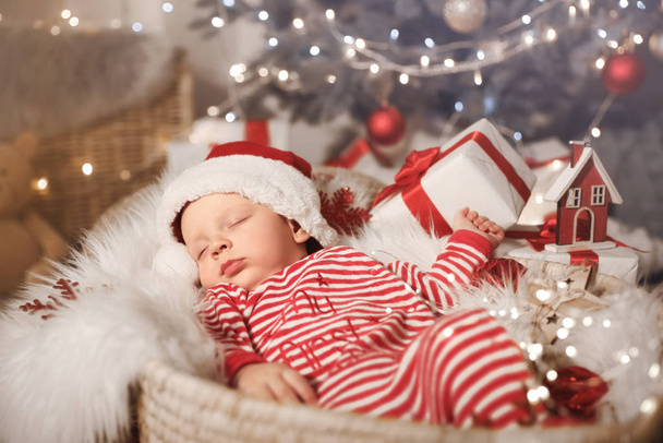 Cute little baby with Santa Claus hat and Christmas gift sleeping in basket at home - Zdjęcie, obraz