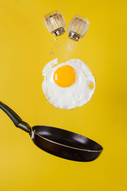 Breakfast time. A fried egg and a frying pan with salt shakers levitate in the air on a yellow background - Photo, Image