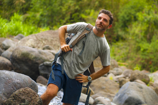 νεαρός ευτυχισμένος και ελκυστικός άντρας με ταξιδιωτικό σακίδιο πεζοπορία στο ποτάμι στο δάσος, απολαμβάνοντας τη φύση και το φρέσκο περιβάλλον στο καλοκαιρινό ταξίδι πεζοπορίας - Φωτογραφία, εικόνα