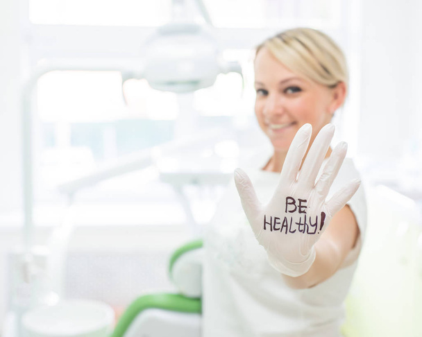 Friendly female dentist smiling and giving high five. Blond doctor in gloves in an office in a medical center. The inscription on the hand be healthy. - Photo, Image