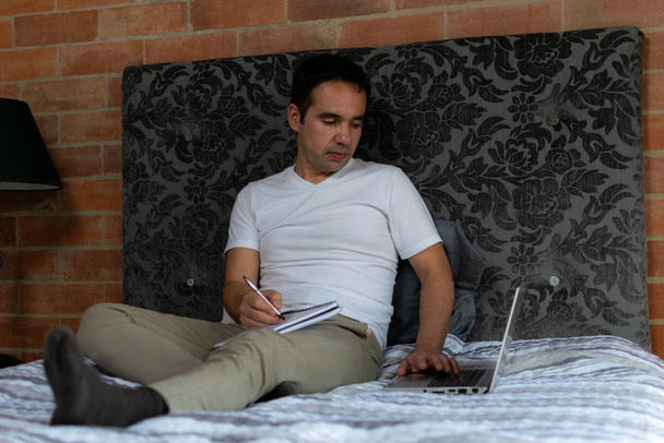 Colombiano latino sentado em sua cama escrevendo em um caderno e verificando seu laptop
 - Foto, Imagem