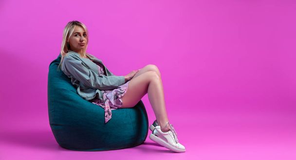 Beautiful blonde girl in a blue jacket sits on a green bag chair - Foto, Imagem