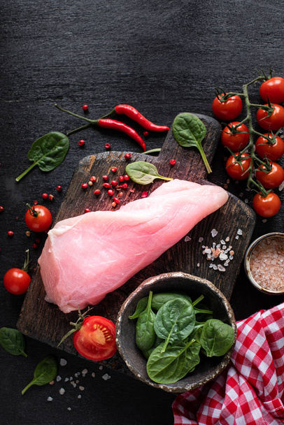 Raw turkey fillet ready for grilling. Chicken fillet on a wooden cutting board with cherry tomatoes, hot pepper, spinach leaves and greens. copy space. top view. Vertical photo - Photo, Image