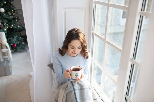 Dark-haired young woman in grey shirt having coffee near the window - Foto, afbeelding