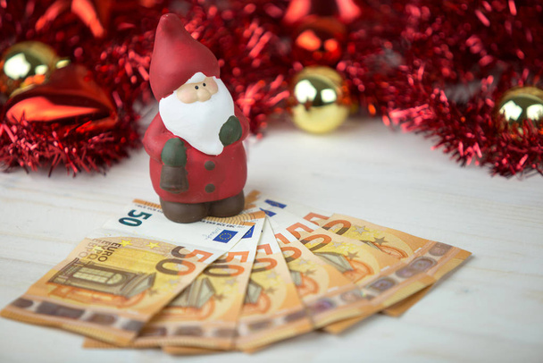 Obchodní koncept vánočních peněz: soška Santa Clause na asi padesáti eurobankovkách s červenými a zlatými cetkami a věnečkovou výzdobou s efektem bokeh na lehkém dřevěném stole - Fotografie, Obrázek