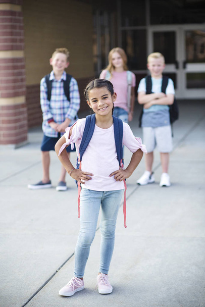Fénykép egy változatos csoport mosolygós általános iskolás diákok előtt áll az iskola épülete. Szelektív fókusz a csinos lány előtt. Kitűnik a tömegből - Fotó, kép