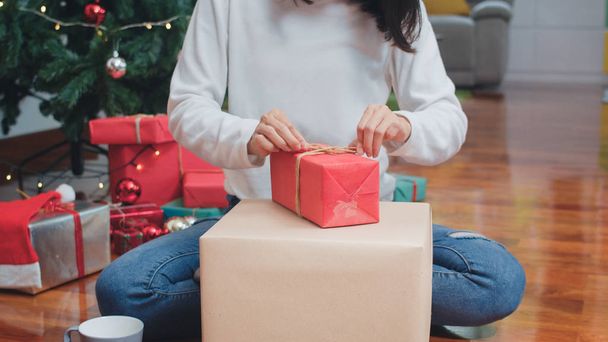 Asian women celebrate Christmas festival. Female teen wear sweater and Christmas hat relax happy wrapping gifts near Christmas tree enjoy xmas winter holidays together in living room at home. - Photo, image