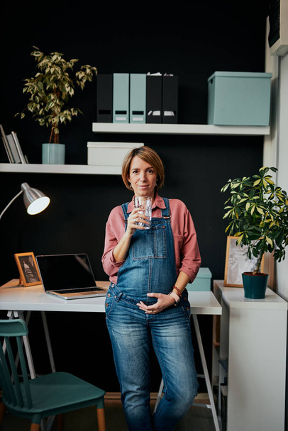 Encantadora mulher grávida caucasiana positiva em pé no escritório em casa, apoiando-se na mesa, segurando vidro de água e tocando barriga
. - Foto, Imagem