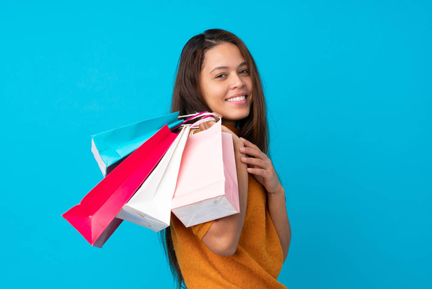 Молодая бразильская девушка на изолированном синем фоне с пакетами покупок и улыбкой
 - Фото, изображение