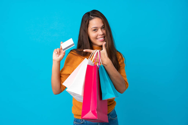 Joven brasileña sobre fondo azul aislado sosteniendo bolsas de compras y una tarjeta de crédito
 - Foto, Imagen