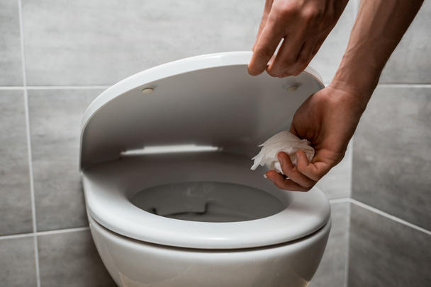 обрізаний вид людини, що тримає кришку туалету і кидає папір в унітаз в сучасному туалеті з сірою плиткою
 - Фото, зображення
