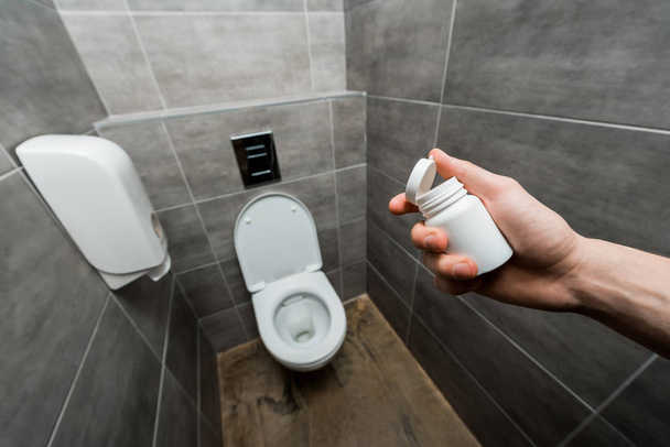 bijgesneden weergave van de man holding container met pillen in de buurt van keramische schoon toilet kom in modern toilet met grijze tegel - Foto, afbeelding