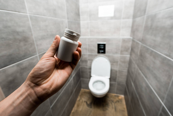 обрезанный вид человека, держащего контейнер с таблетками рядом с керамической чистой унитаз в современном туалете с серой плиткой
 - Фото, изображение