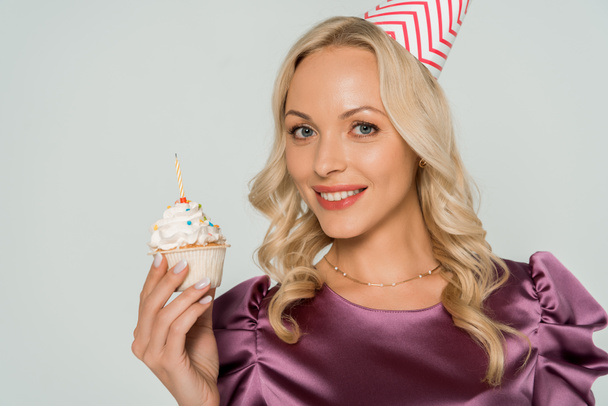 привлекательная, веселая женщина в кепке для вечеринок с праздничным кексом и смотрящая на камеру, изолированную на сером
 - Фото, изображение