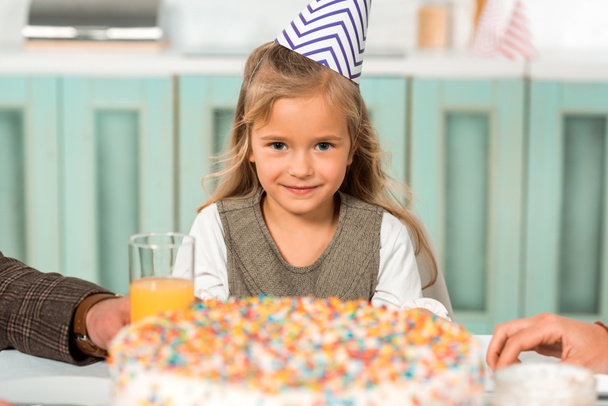 επιλεκτική εστίαση του αξιολάτρευτο παιδί σε κόμμα καπάκι κοιτάζοντας κάμερα, ενώ κάθεται κοντά τούρτα γενεθλίων - Φωτογραφία, εικόνα