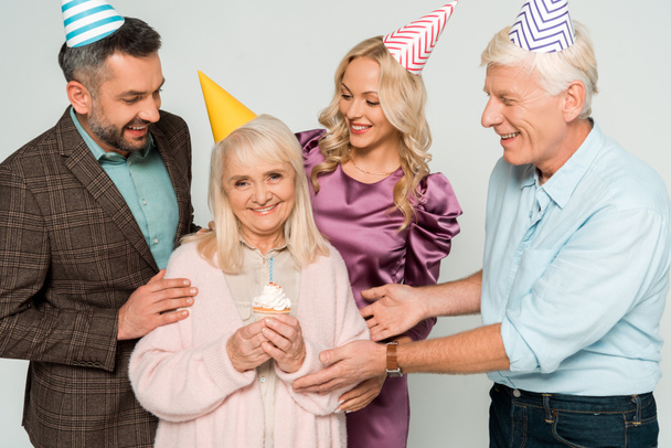 happy senior woman holding birthday cupcake near husband and adult daughter and son isolated on grey - Foto, afbeelding