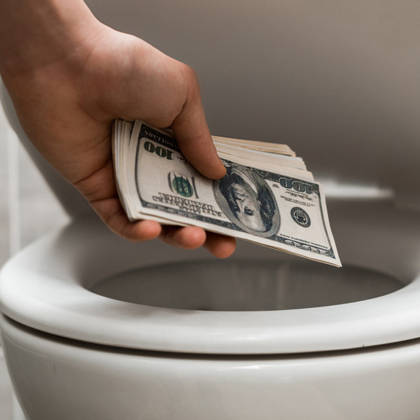 обрезанный вид человека с долларовыми банкнотами возле унитаза в современном туалете с серой плиткой
 - Фото, изображение