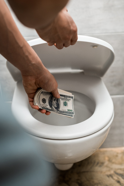 bijgesneden beeld van de mens gooien dollarbankbiljetten in toilet kom - Foto, afbeelding