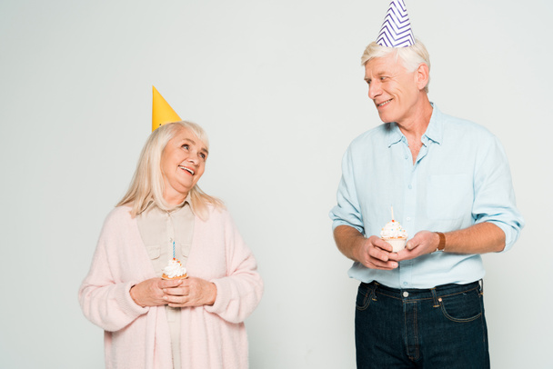 happy senior husband and wife holding birthday cupcakes and looking at each other isolated on grey - Fotoğraf, Görsel