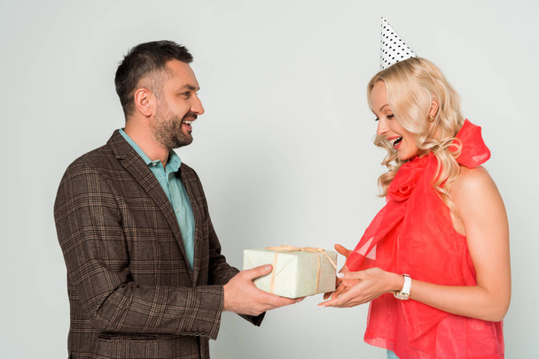 smiling man presenting gift box to happy wife on grey background - Photo, image