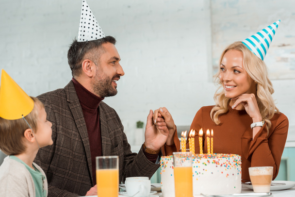 happy man and woman holding hands while sitting at table with son near birthday cake with burning candles - Zdjęcie, obraz