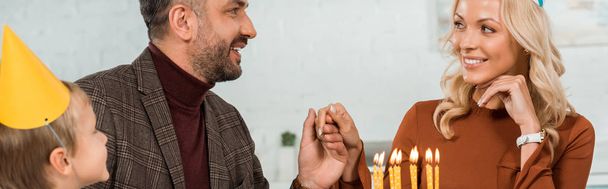 panoramic shot of happy man and woman holding hands while sitting at table with son near birthday cake with burning candles - Photo, Image