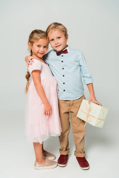 cute boy holding gift box and embracing adorable sister on grey background - Photo, image