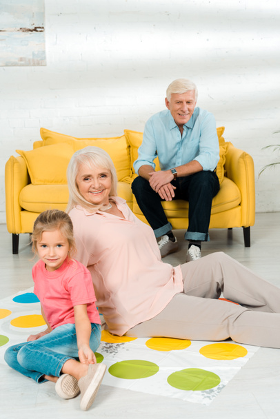 KYIV, UKRAINE - NOVEMBER 21, 2019: senior woman sitting on twister game mat with granddaughter near husband sitting on sofa - Foto, Imagem