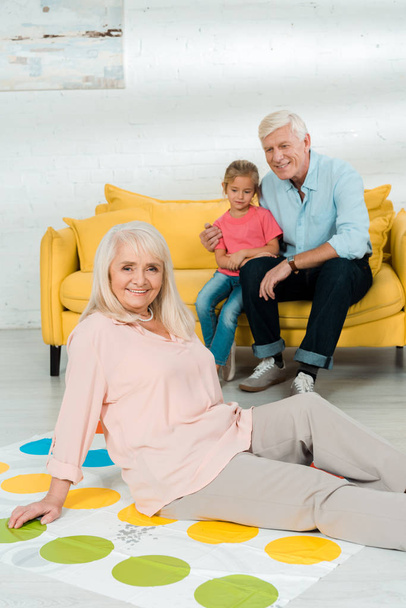 KYIV, UKRAINE - NOVEMBER 21, 2019: smiling senior woman sitting on twister game mat near husband and grandson sitting on sofa - Zdjęcie, obraz