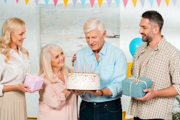 šťastný senior muž drží narozeninový dort, zatímco stojí v blízkosti rodiny - Fotografie, Obrázek