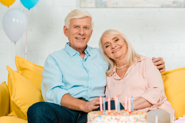 happy senior husband and wife sitting near birthday cake and smiling at camera - Photo, Image
