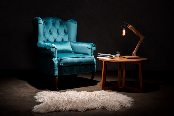 элегантное велюровое синее кресло с подушкой рядом с ковровым покрытием животного, деревянный стол и лампа изолированы на черном
 - Фото, изображение