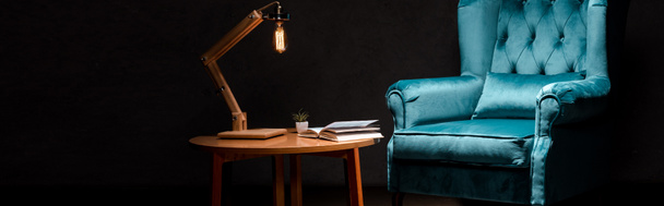 elegant velour blue armchair with pillow near wooden table and lamp isolated on black, panoramic shot - Foto, afbeelding
