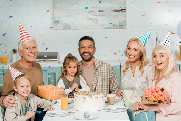 onnellinen perhe istuu keittiön pöydässä lähellä syntymäpäiväkakkua putoavan konftin alla
 - Valokuva, kuva