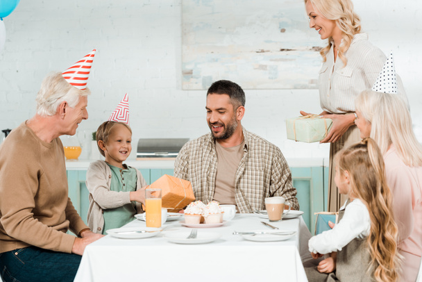 笑顔の女性と子供は家族の近くのキッチンテーブルに座って幸せな父親に贈り物を提示します - 写真・画像