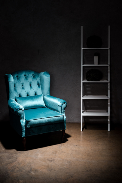 κομψή βελούδινη μπλε πολυθρόνα με μαξιλάρι κοντά στο ράφι στο σκοτεινό δωμάτιο - Φωτογραφία, εικόνα