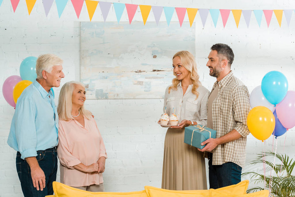 smiling man and woman holding birthday cupcakes and gift box while looking at senior parents - Фото, изображение