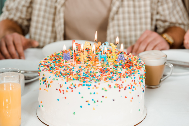 bijgesneden uitzicht op de mens zitten in de buurt van verjaardagstaart met kleurrijke brandende kaarsen - Foto, afbeelding