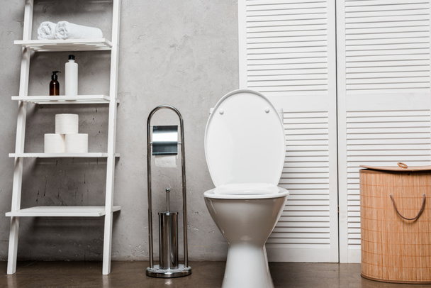 intérieur de la salle de bain moderne avec cuvette de toilette près de rack avec cosmétiques, serviettes, papier toilette, panier à linge et brosse de toilette
 - Photo, image