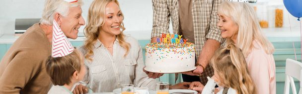 vista recortada del hombre sosteniendo pastel de cumpleaños con velas cerca de la familia feliz sentado en la mesa de la cocina
 - Foto, Imagen