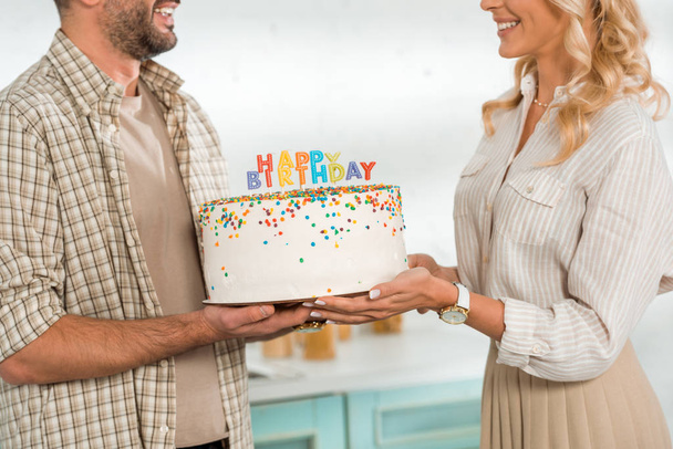 cropped view of smiling husband and wife holding birthday cake with colorful candles and happy birthday lettering - Foto, immagini