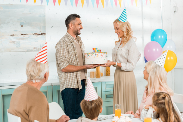 усміхнений чоловік презентує торт на день народження щасливій дружині, поки вся сім'я сидить за кухонним столом
 - Фото, зображення